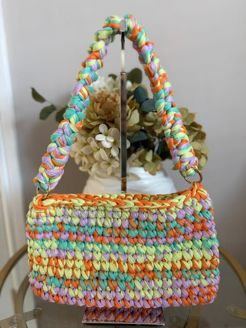 A handmade multicolor crochet handbag with braided shoulder straps, displayed on a stand with dried flowers in the background.