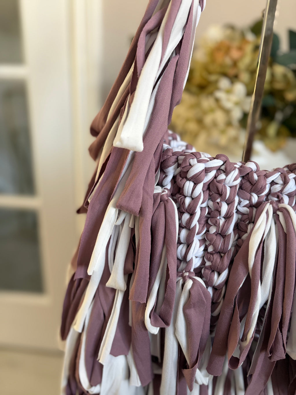A close-up of a crochet bag with long fringe straps in dusty mauve and white, 