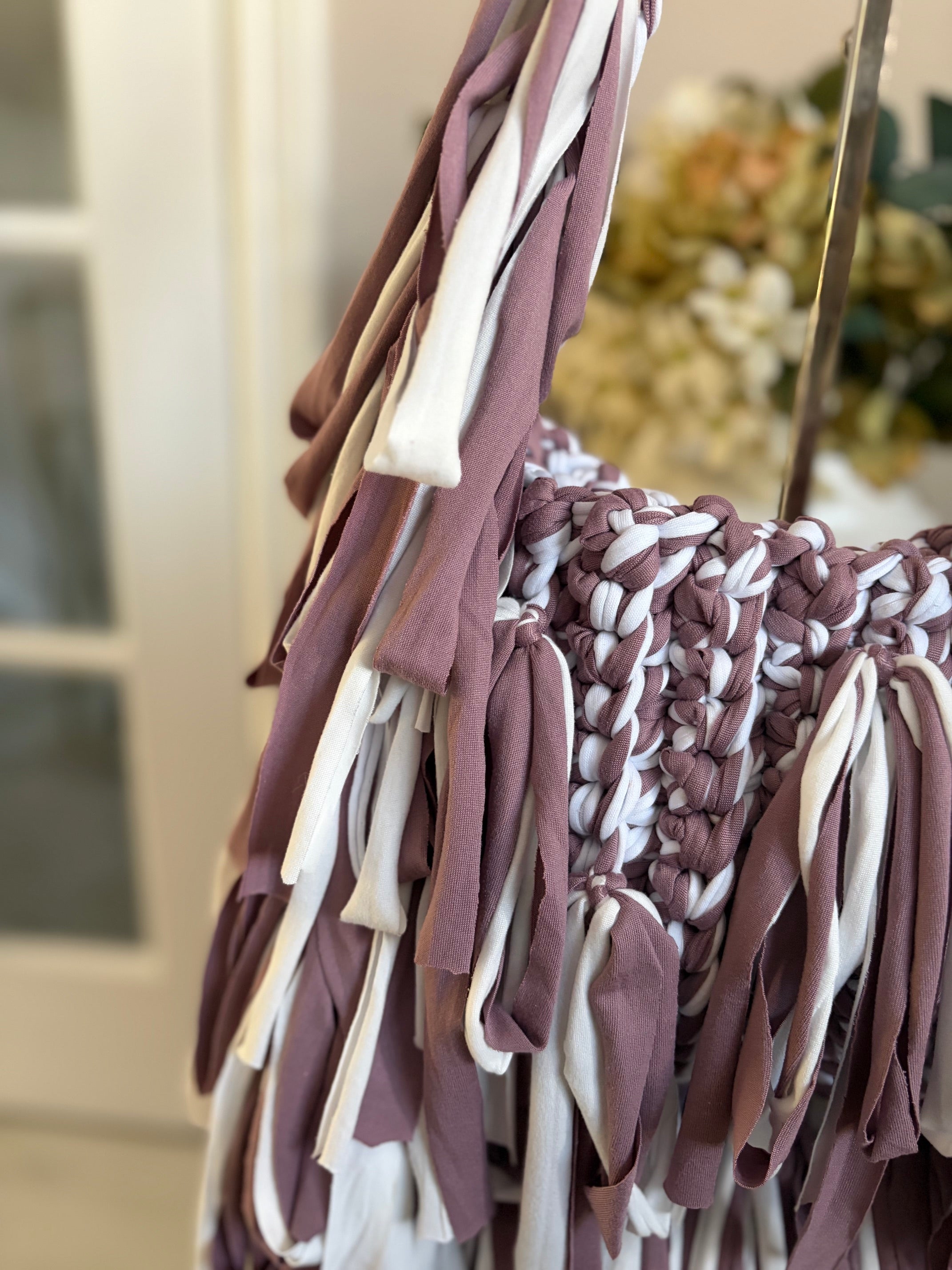 A close-up of a crochet bag with long fringe straps in dusty mauve and white, 