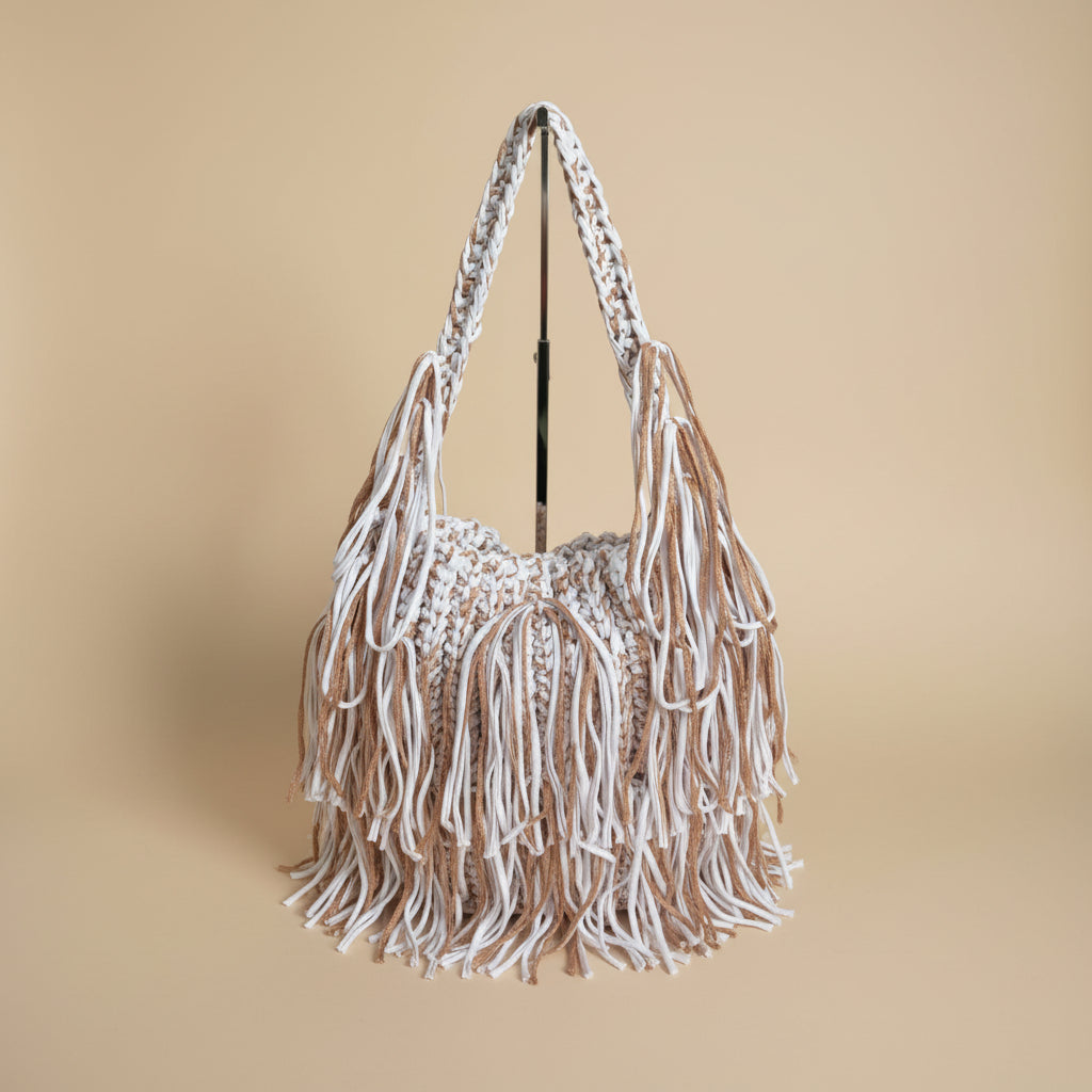 A close-up of a crochet bag with long fringe handles and tassels, set against a softly blurred indoor background with a vase of flowers.