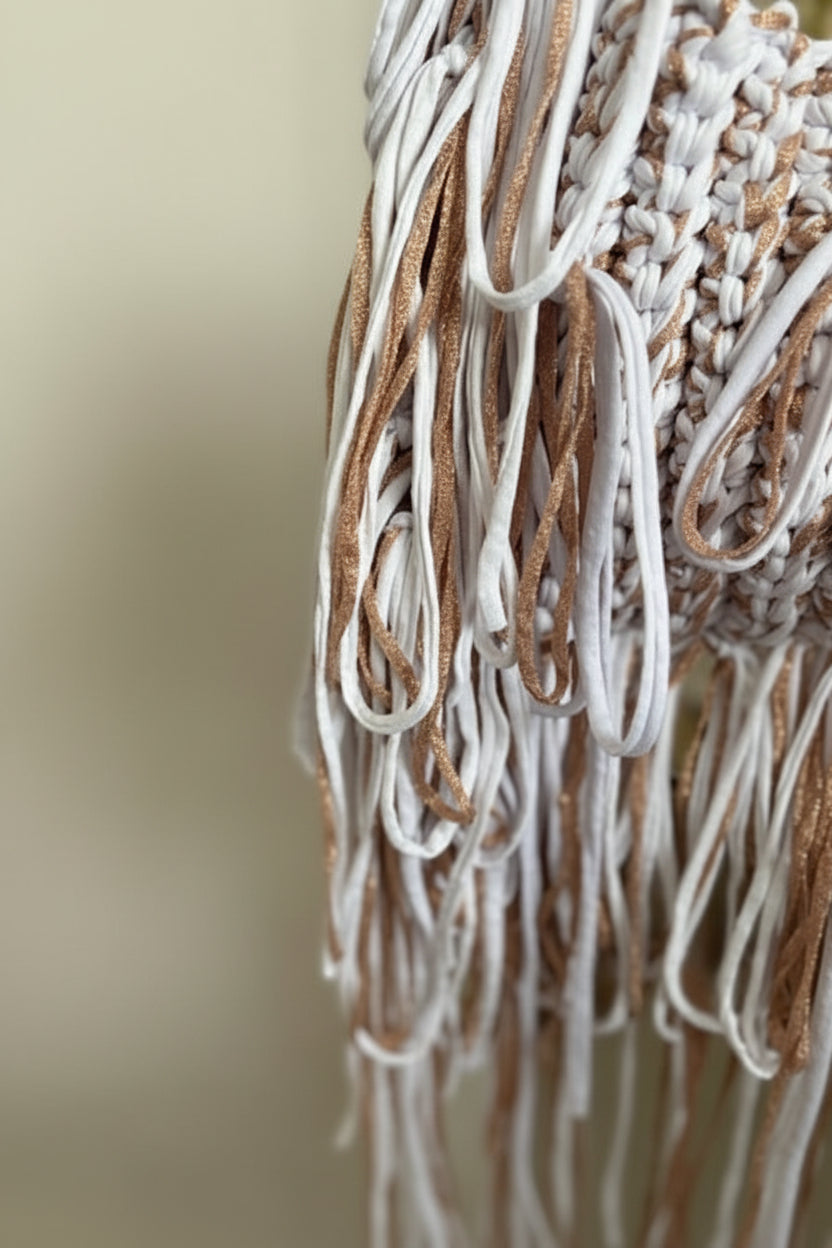 Close-up of a woven textile with long fringe in white and metallic gold tones, shown against a softly blurred indoor background.