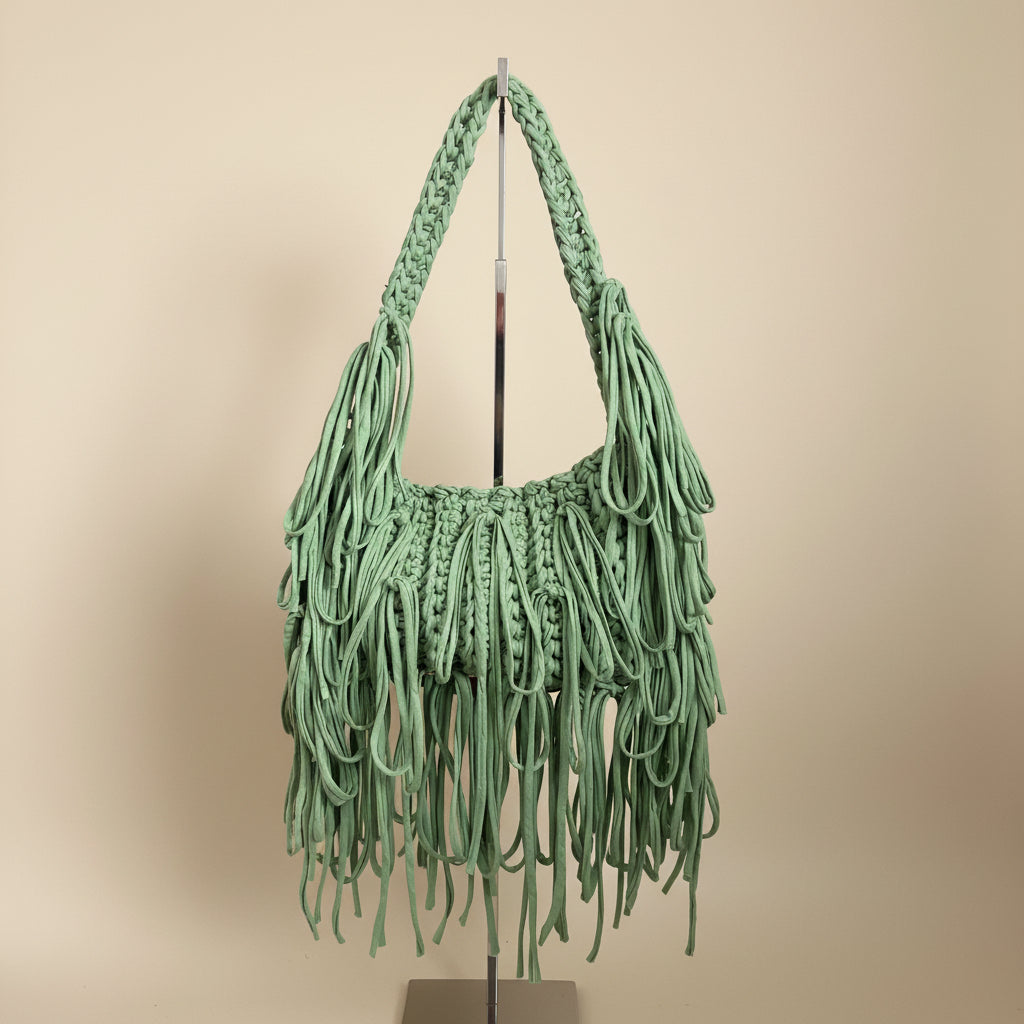 A sage green crochet shoulder bag with long fringed tassels and a thick braided strap