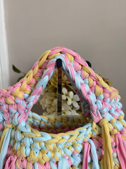 A close-up of a handmade braided tote bag made from thick pastel yarn in pink, light blue, and yellow.