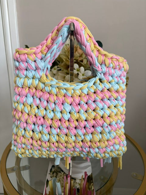 A chunky handwoven tote bag made from thick braided yarn in pastel pink, light blue, and pale yellow, displayed on a glass-topped table.