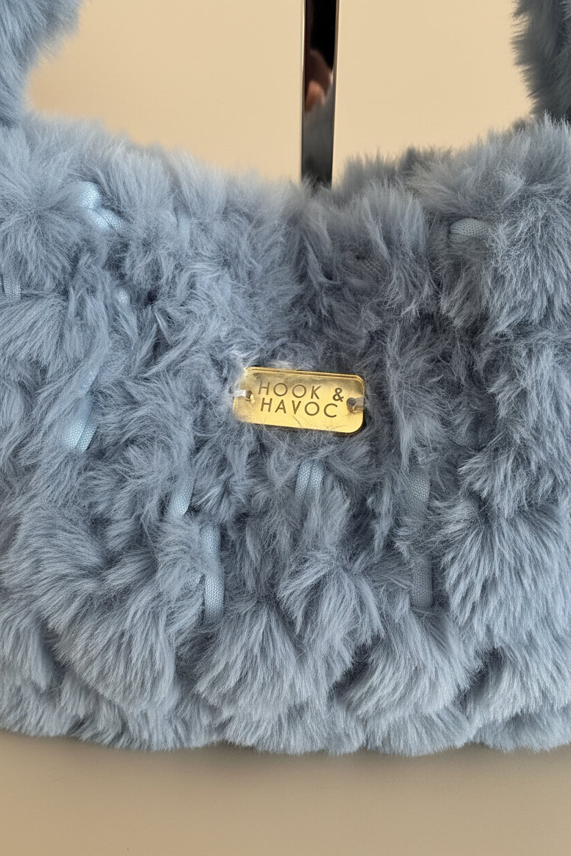 Close-up of a fluffy light blue-gray faux fur textile with a small gold metal tag reading 'HOOK & HAVOC'.