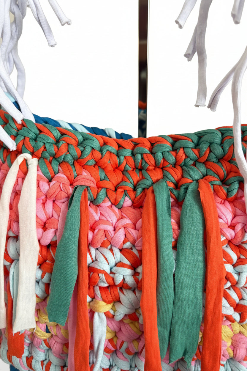 Close-up of a colourful crochet bag with orange, teal, pink, white, and light blue strips, alongside white fringe details.