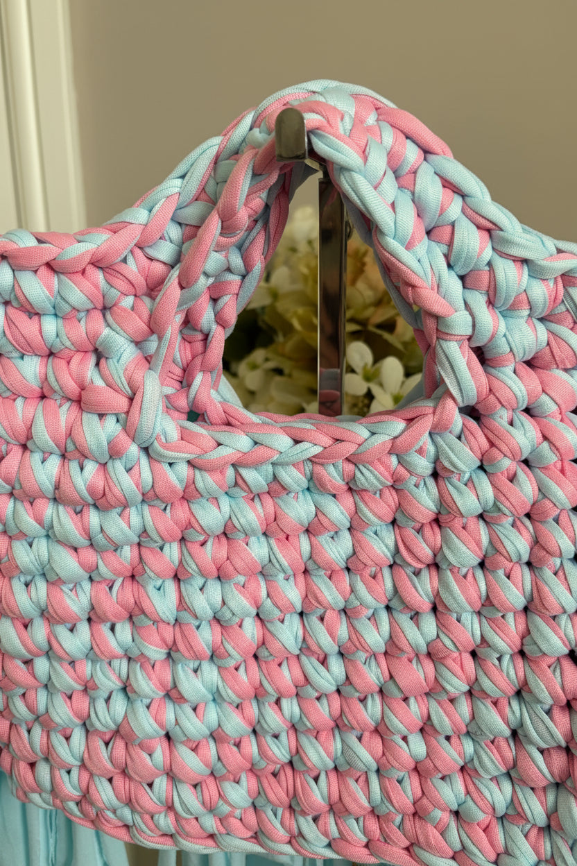 A close-up of a chunky handwoven bag made from thick braided yarn in alternating pastel pink and light blue
